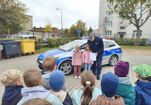 Dwie dziewczynki stoją przy policjancie, który trzyma podziękowanie. Za nimi samochód policyjny.