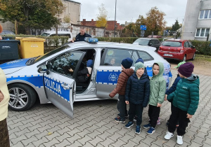 Samochód policyjny z otwartymi drzwiami, obok stoją chłopcy. Z drugiej strony samochodu stoi policjant.