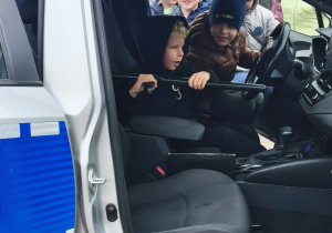 Chłopiec siedzi w samochodzie policyjnym, trzyma pałkę policyjną. Za nim wchodzi do samochodu następny chłopiec.