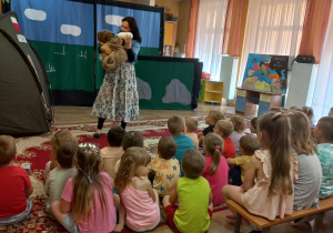 Dzieci siedzą na dywanie i ławeczce. Oglądają spektakl. Aktorka karmi maskotkę - misia mlekiem z butelki.