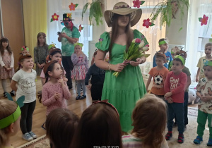 Przedszkolaki a wśród nich kobieta przebrana za Wiosnę w zielonej sukni i słomkowym kapeluszu.