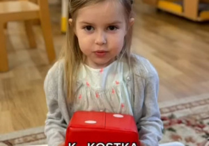 Dziewczynka trzyma czerwoną kostkę do zabawy z białymi kropkami. Napis :K-Kostka.