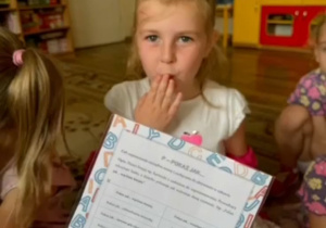 Dziewczynka zakrywa usta dłonią. Napis: P-Pokaż jak...