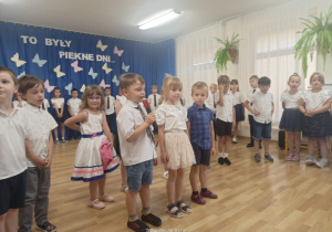 Dwóch chłopców i dziewczynka mówią do mikrofonu za nimi grupa przedszkolaków.