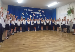Przedszkolaki stoją w półokręgu, trzymają się za ręce wysoko uniesione. Za nimi na granatowym tle kolorowe, papierowe motyle i napis: To były piękne dni...