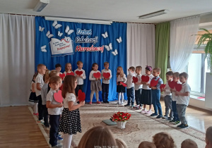 Dzieci 6-letnie, odświętnie ubrane, stoją w półokręgu trzymają czerwone serca, na środku stoi biały kosz z kwiatami w kształcie serc. Na dekoracji napis: Dzień Edukacji Narodowej poniżej otwarta księga i napis: za trud, za uśmiech, za serce. Dziękujemy!