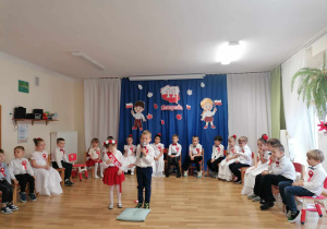 Natalka z Julkiem z grupy Żuczkó recytują swoje wiersze. W tle w półkolu ubrane na biało-czerwono Żuczki występują na tle pieknej, tematycznej dekoracji.
