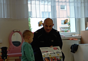 Pan policjant trzyma planszę z numerami alarmowymi, obok stoi chłopiec i uważnie przygląda się planszy.