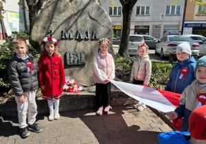 Dzieci stoją pod pomnikiem z rozłożoną flagą.