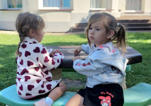 Dwie dziewczynki siedzą przy ogrodowym stoliku. w tle budynek przedszkola.