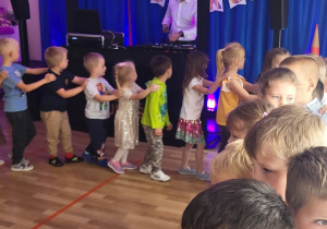 Dzieci w tanecznym wężu. Na granatowym tle kolorowy napis Dzień Przedszkolaka.