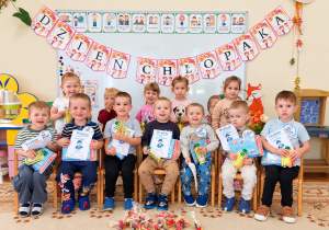 Chłopcy siedzą na krzesłach, każdy trzyma dyplom, książkę i soczek. Za nimi stoją dziewczynki. U góry napis Dzień Chłopaka.