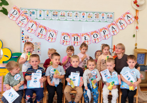 Chłopcy siedzą na krzesłach, każdy trzyma dyplom, książkę i soczek. Za nimi stoją dziewczynki i nauczycielka. U góry napis Dzień Chłopaka
