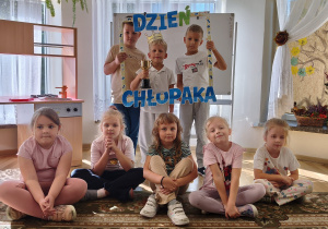 Dwóch chłopców trzyma ramkę z napisem Dzień Chłopaka, w niej chłopiec w koronie trzyma puchar. Przed chłopcami siedzi pięć dziewczynek.