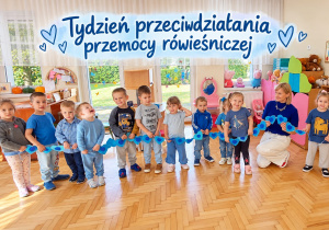 Maluszki i nauczycielka, ubrani na niebiesko, trzymają łańcuch z niebieskich serc. Nad nimi napis: Tydzień przeciwdziałania przemocy rówieśniczej.