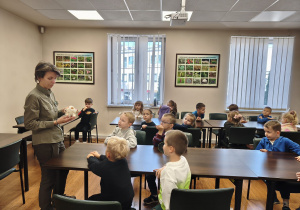 Dzieci siedzą przy stolikach. Pani leśnik trzyma czaszkę zwierzęcia. Na ścianach dwie tablice z obrazkami roślin.