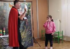 Iluzjonista pokazuje dziewczynce w różowej bluzie kartę. Dziecko zakrywa usta dłonią.