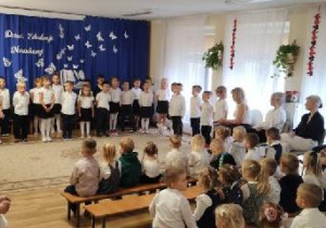 Dzieci w odświętnych strojach stoją w półkolu, za nimi dekoracja na granatowym tle napis Dzień Edukacji Narodowej, biała księga, z której wylatują białe sylwety motyli.