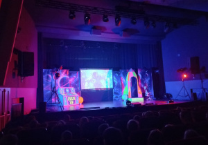 Scena, na której stoi dekoracja Kosmicznego Show: rakieta, konsola z przyciskami, ekran interaktywny. Na widowni siedzą dzieci.