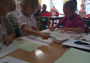 Dzieci w sali przedszkolnej siedzą przy stoliku i wykonują zielnik.