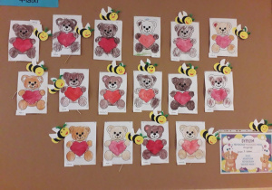 Tablica z pracami dzieci 4-letnich: miś trzyma czerwone serce-kolorowanka, pszczółki na patykach. Dyplom Przyjaciela Pluszowego misia dla dzieci z grupy 4-latków.