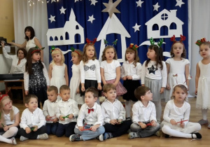 Dzieci z grupy 4-,5-latków śpiewają i akompaniują na patyczkach pastorałkę za nimi świąteczna dekoracja.