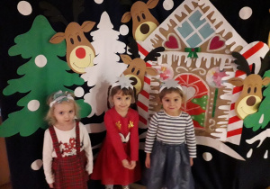 Trzy dziewczynki z grupy 3-latków na tle zimowej dekoracji: las, renifery i domek z piernika.