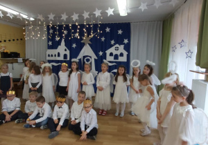 Dzieci sześcioletnie odświętnie ubrane, śpiewają pastorałkę podczas Wigilii w przedszkolu na tle bożonarodzeniowej dekoracji.