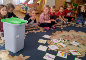 Dzieci siedzą na dywanie. Przed nimi leżą obrazki z narysowanymi odpadami do posegregowania. Na podłodze stoi kosz z zieloną pokrywą.