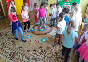Przedszkolaki poruszają się pomiędzy ułożonymi na dywanie papierowymi kwiatami.