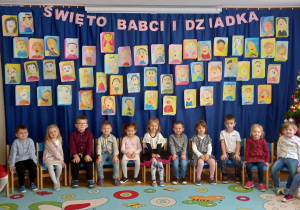 Dzieci 4-letnie siedzą w półokręgu na krzesełkach. W tle portrety Babć i Dziadziusiów narysowane przez przedszkolaków.