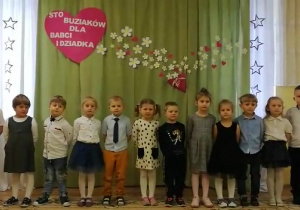 Dzieci z grupy Słoneczka przygotowują się do śpiewu piosenki dla babci i Dziadka. W tle serce z napisem "Sto buziaków dla babci i Dziadka"..