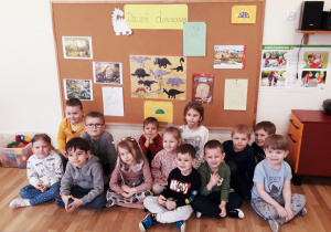 Dzieci 4- i 5-letnie siedzą przed tablicą, na której jest napis "Dzień dinozaura". Pod napisem umieszczone są rysunki i ilustracje dinozaurów.