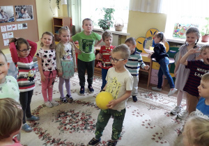 Dzieci, w sali przedszkolnej, stoją w kole, na środku Mikołaj z żółtą piłką.