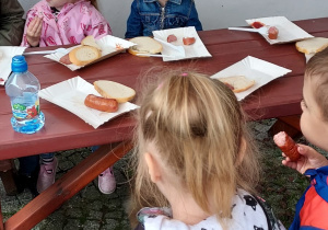 Dzieci siedzą przy stoliku i jedzą pieczone kiełbaski.