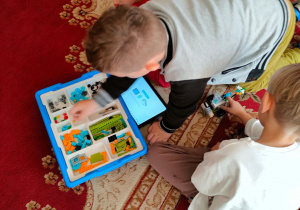 Chłopcy budują na dywanie roboty z klocków lego, według instrukcji podanej na tablecie.
