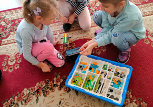 Dziewczynki budują na dywanie roboty z klocków lego, według instrukcji podanej na tablecie.