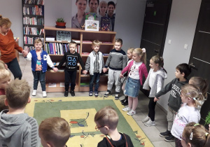 Przedszkolaki trzymają się za ręce, stoją w kręgu w bibliotecznej sali.