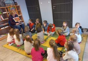Dzieci siedzą na dywanie i słuchają bajkę czytaną przez panią bibliotekarkę.