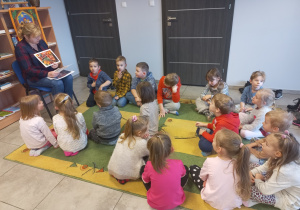 Dzieci siedzą na dywanie i słuchają bajkę czytaną przez panią bibliotekarkę.
