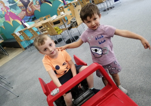 Chłopiec siedzi na dużym zabawkowym samochodzie, drugi chłopiec trzyma go za ramię.