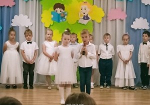 Dziewczynka i chłopiec stoją pośrodku sali. Chłopiec trzyma mikrofon i recytuje wiersz. Za nimi w rzędzie stoją dzieci.