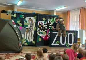 Sceneria przedstawiająca egzotyczne zwierzęta, napis ZOO. Aktor żołnierz obok niego duży pluszowy niedźwiedź Wojtek.