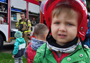 Chłopiec w kasku strażackim. Za nim wóz strażacki i strażak.