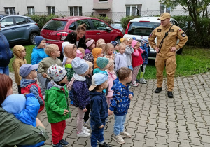 Przedszkolaki i nauczyciele stoją przed wozem strażackim i słuchają ciekawostek o pracy strażaków, o których opowiada pan strażak.