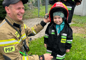 Chłopiec ubrany w strój strażaka i dwóch przedstawicieli Państwowej Straży Pożarnej.