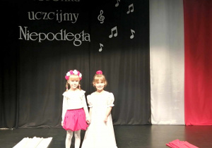 Amelka i Marysia w pięknych, biało-czerwonych strojach na tle napisu Piosenką uczcijmy Niepodległą.