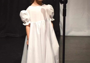 Marysia w pięknej, białej sukience z czerwonym kwiatem we włosach, spiewa na scenie konkursową piosenkę.