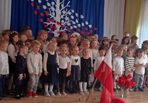 Przedszkolaki stoją na tle biało-czerwonego drzewa i śpiewają hymn Polski. Przed nimi stoją w wazonie czerwone kwiaty i flaga Polski.