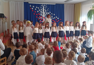 Dzieci z grupy sześciolatków podczas inscenizacji z okazji Święta Niepodległości, za nimi pani Ewa.
